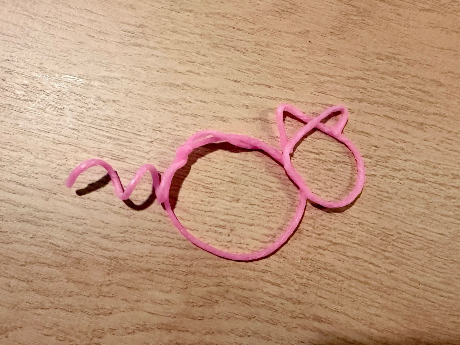 A pig made from a pink Wikki Stix: a round body, a smaller round head with two pointed ears, and a curly tail, lying on a wooden table.